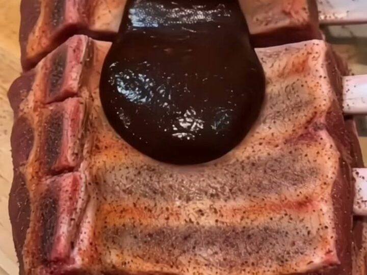 A close-up shot of glossy, dark barbecue sauce being ladled over tender, slow-cooked ribs on a wooden cutting board.