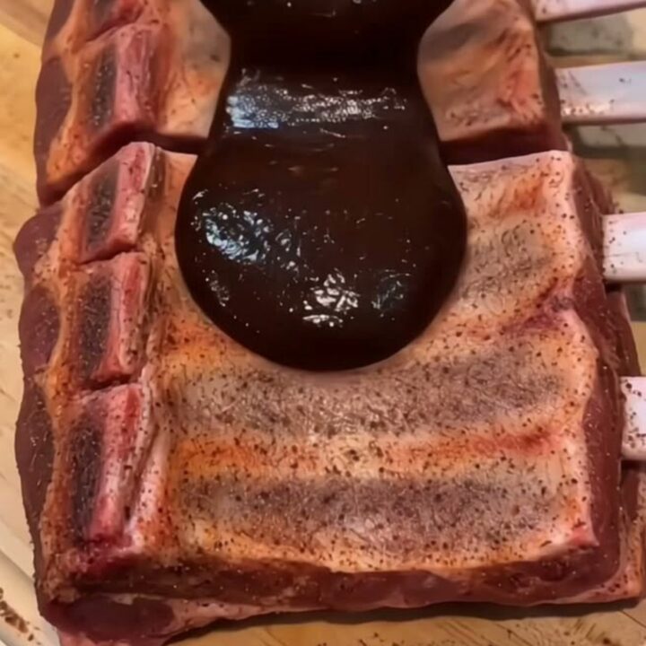 A close-up shot of glossy, dark barbecue sauce being ladled over tender, slow-cooked ribs on a wooden cutting board.