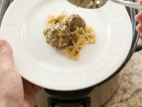 Slow cooker filled with creamy meatball pesto farfalle, topped with melted mozzarella.