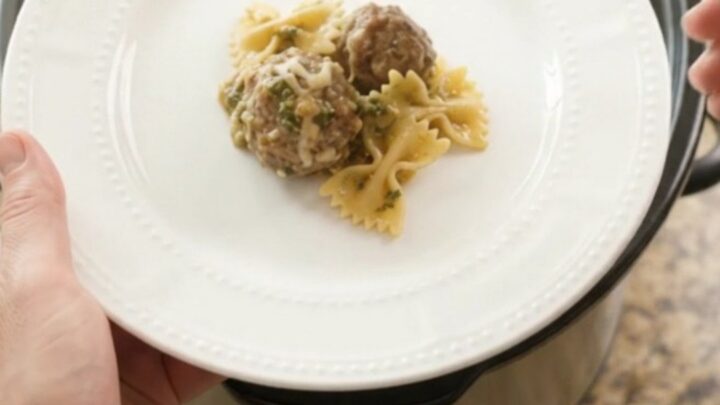 Slow cooker filled with creamy meatball pesto farfalle, topped with melted mozzarella.