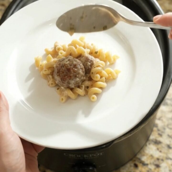 Slow cooker filled with creamy cavatappi pasta, meatballs, and melted provolone cheese, with a spoon lifting a cheesy scoop.