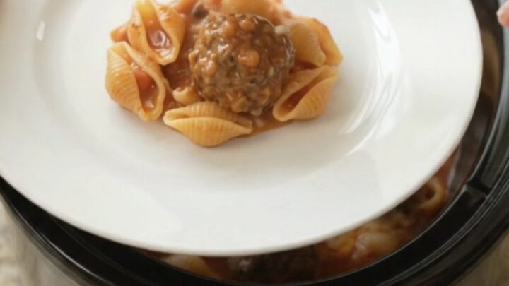 Slow cooker filled with meatballs, pasta shells, and red pepper sauce topped with ricotta and provolone.