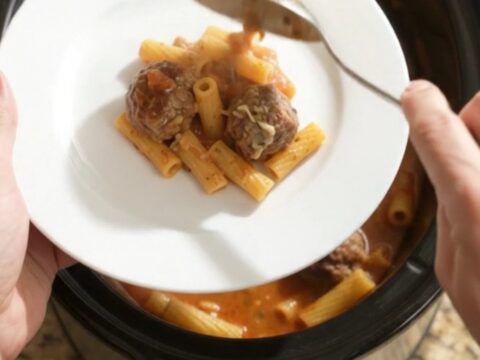 A slow cooker filled with bubbling Boursin meatball baked ziti, with cheese stretched from a spoon.