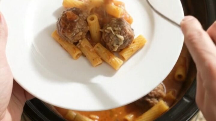 A slow cooker filled with bubbling Boursin meatball baked ziti, with cheese stretched from a spoon.