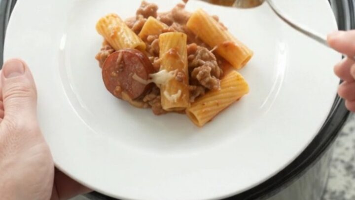 Slow cooker full of cheesy rigatoni with kielbasa and tomato sauce, golden crust on top.
