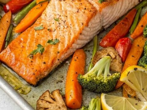 A sheet pan filled with roasted salmon fillets, broccoli florets, and sliced carrots with lemon wedges.