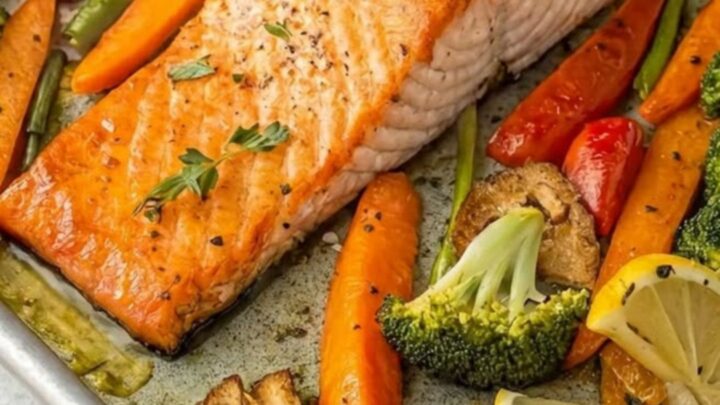 A sheet pan filled with roasted salmon fillets, broccoli florets, and sliced carrots with lemon wedges.