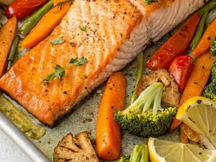 A sheet pan filled with roasted salmon fillets, broccoli florets, and sliced carrots with lemon wedges.