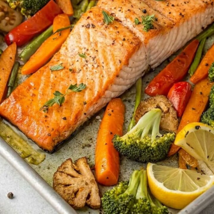 A sheet pan filled with roasted salmon fillets, broccoli florets, and sliced carrots with lemon wedges.