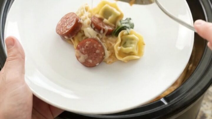 Slow cooker filled with creamy kielbasa tortellini Alfredo, with melted cheese and mushrooms visible