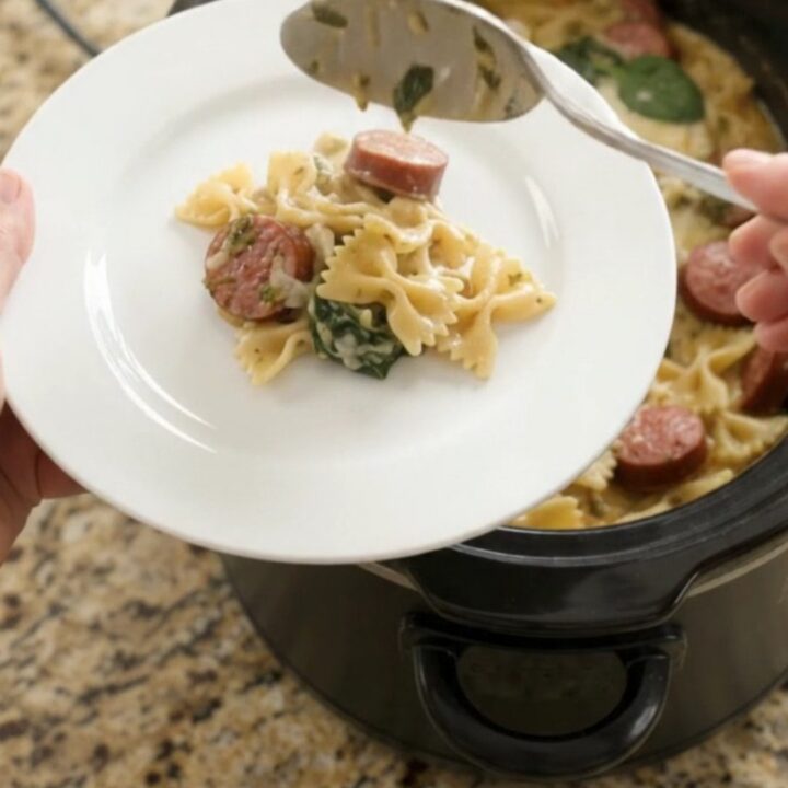 Creamy slow-cooker farfalle with smoked kielbasa, spinach, and melted cheese in a black crockpot
