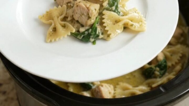 Slow cooker filled with creamy Parmesan chicken pesto farfalle, with melted cheese and wilted spinach