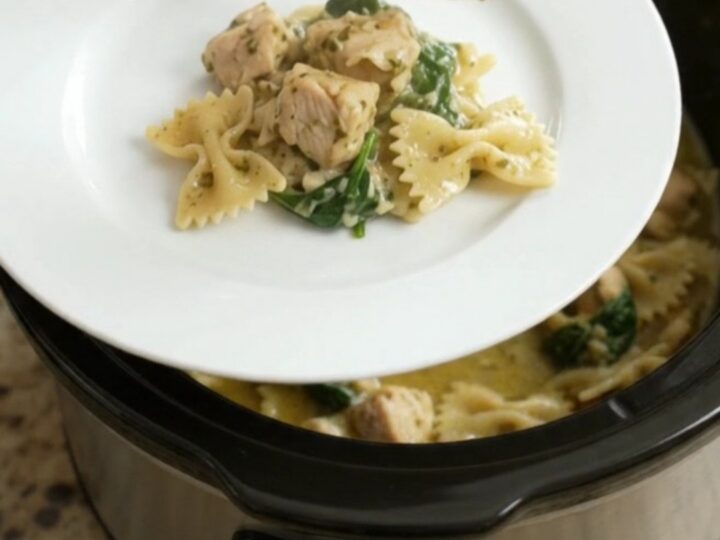 Slow cooker filled with creamy Parmesan chicken pesto farfalle, with melted cheese and wilted spinach