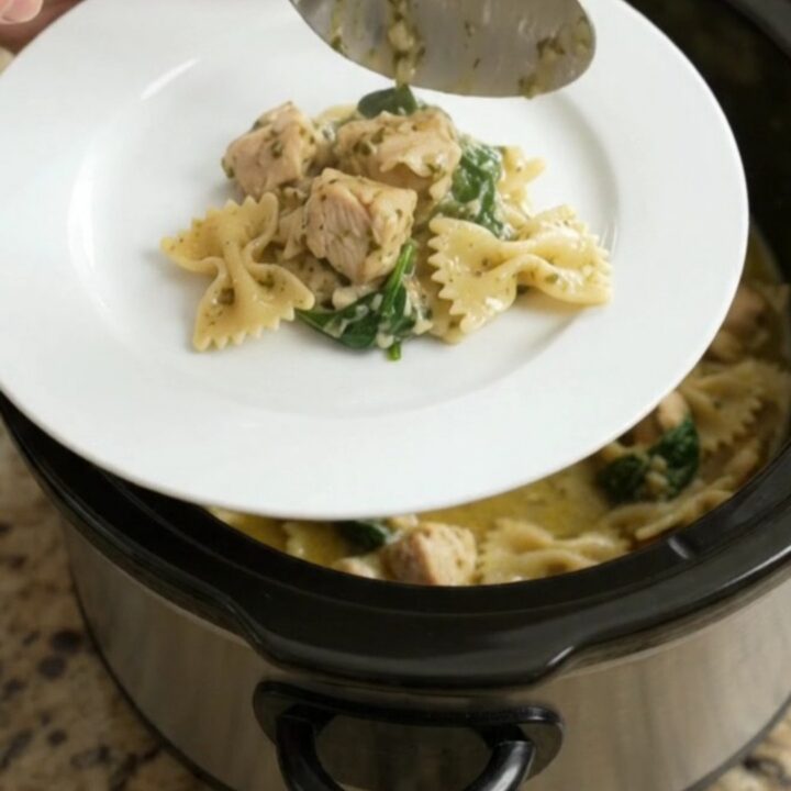 Slow cooker filled with creamy Parmesan chicken pesto farfalle, with melted cheese and wilted spinach