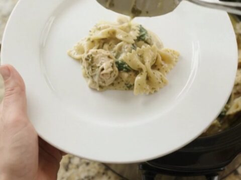 Slow cooker filled with creamy chicken pesto farfalle, topped with melted cheese and spinach