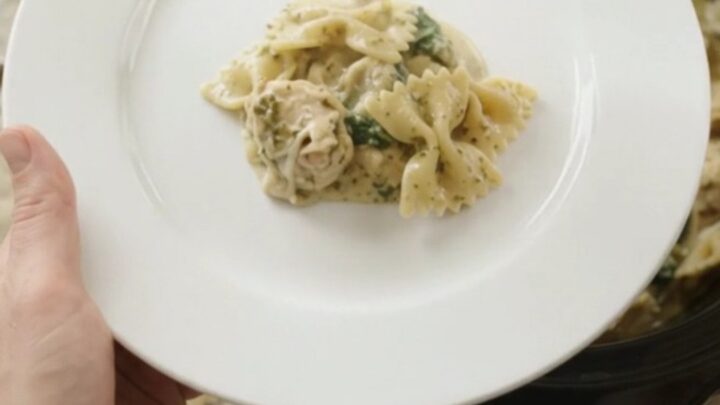 Slow cooker filled with creamy chicken pesto farfalle, topped with melted cheese and spinach