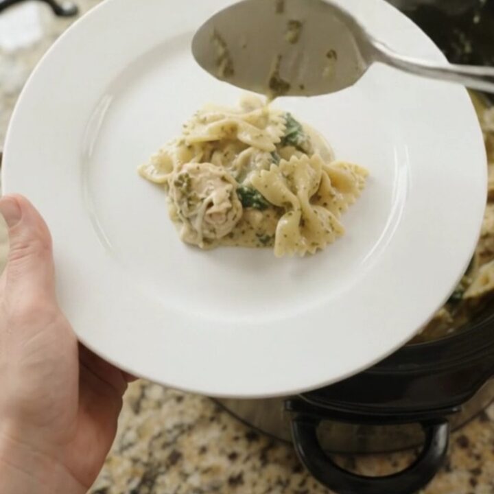 Slow cooker filled with creamy chicken pesto farfalle, topped with melted cheese and spinach