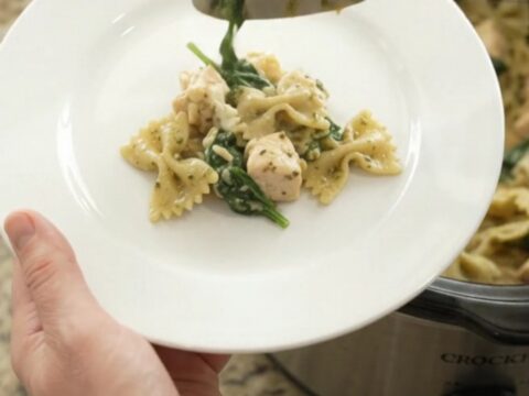 A slow cooker filled with creamy chicken pesto farfalle, topped with melted provolone and fresh basil.