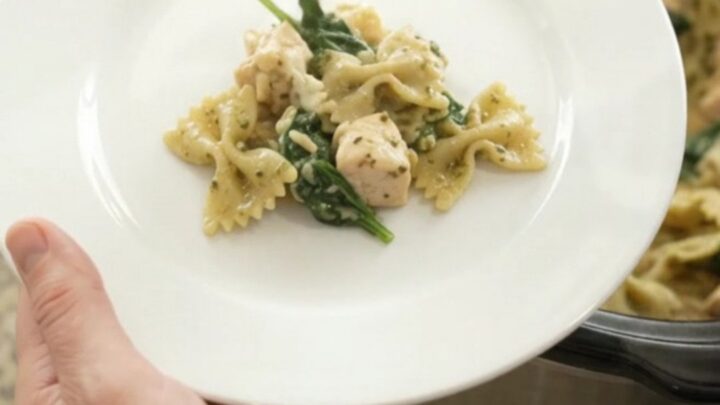 A slow cooker filled with creamy chicken pesto farfalle, topped with melted provolone and fresh basil.