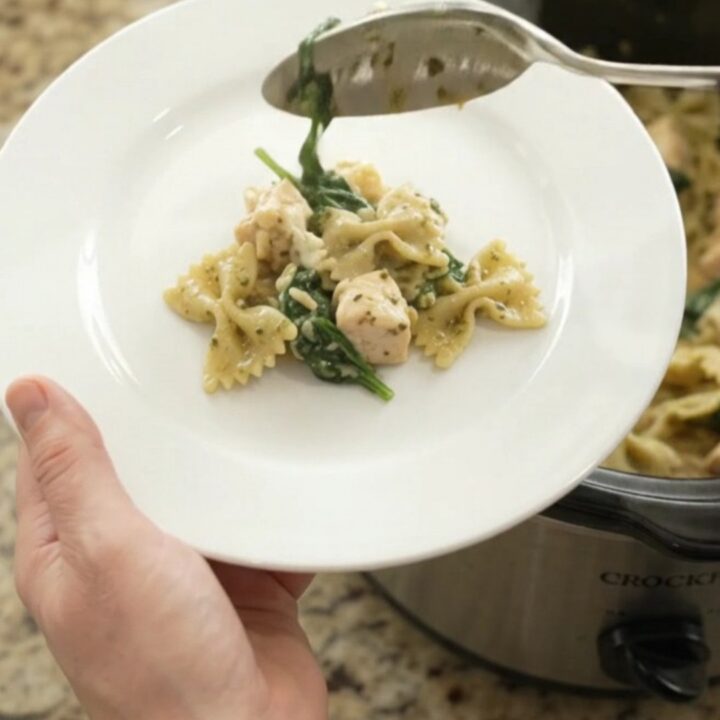 A slow cooker filled with creamy chicken pesto farfalle, topped with melted provolone and fresh basil.