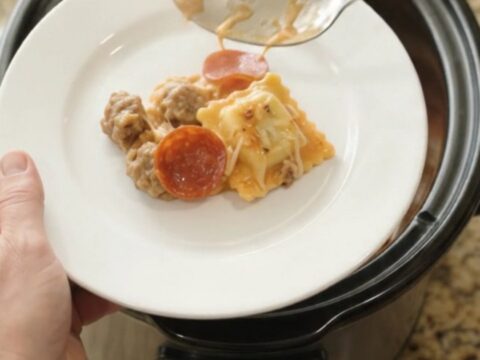 A slow cooker filled with cheesy pepperoni sausage vodka ravioli, with a spoon lifting a stretchy, steaming portion.