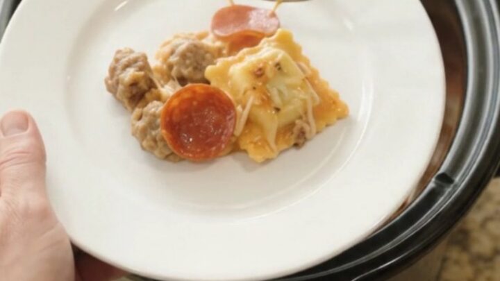 A slow cooker filled with cheesy pepperoni sausage vodka ravioli, with a spoon lifting a stretchy, steaming portion.