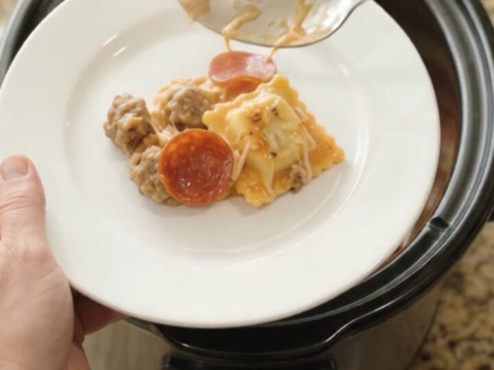 A slow cooker filled with cheesy pepperoni sausage vodka ravioli, with a spoon lifting a stretchy, steaming portion.