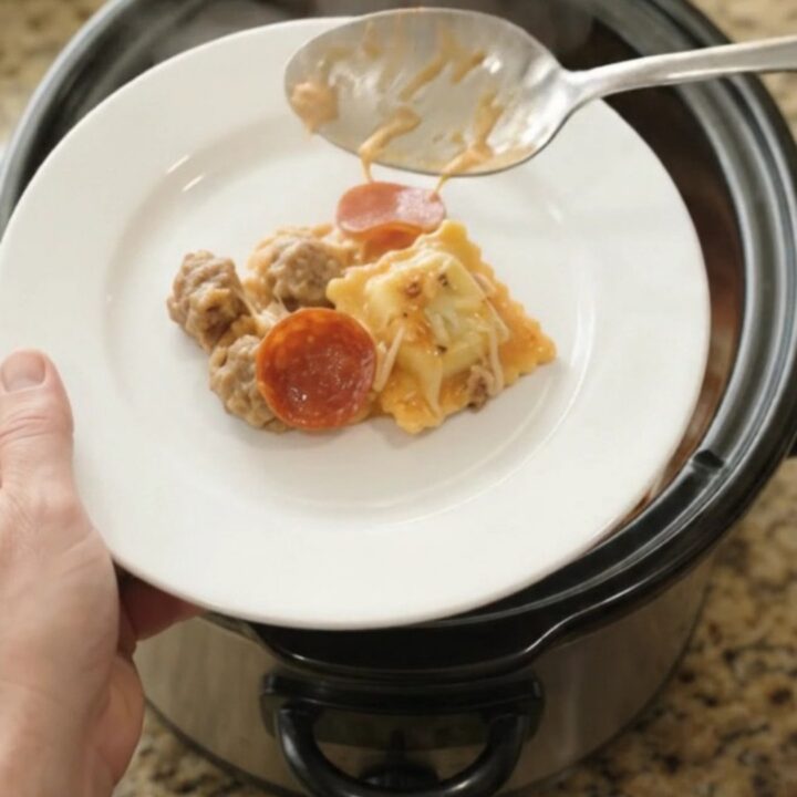 A slow cooker filled with cheesy pepperoni sausage vodka ravioli, with a spoon lifting a stretchy, steaming portion.