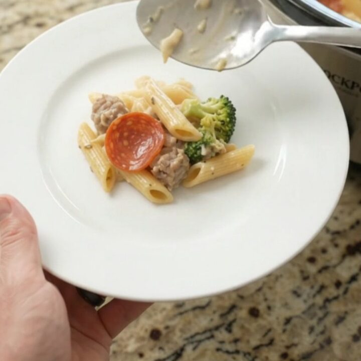 Slow cooker filled with creamy penne pasta, Italian sausage, pepperoni, and broccoli in Alfredo sauce with melted mozzarella