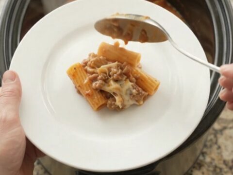 A slow cooker filled with bubbling rigatoni, pepperoni, sausage, and melted cheese, with steam rising.