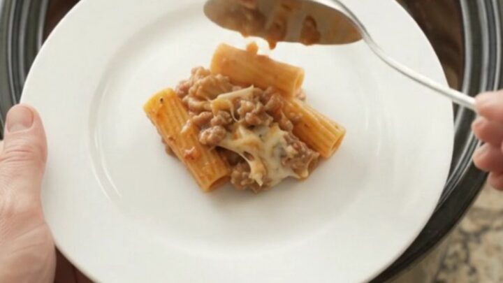 A slow cooker filled with bubbling rigatoni, pepperoni, sausage, and melted cheese, with steam rising.