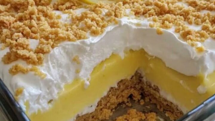 A tall, creamy slice of lemon dessert in a glass dish showing distinct layers of crust, pudding, and whipped cream.
