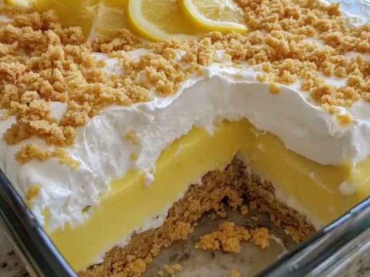 A tall, creamy slice of lemon dessert in a glass dish showing distinct layers of crust, pudding, and whipped cream.