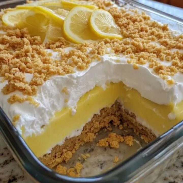 A tall, creamy slice of lemon dessert in a glass dish showing distinct layers of crust, pudding, and whipped cream.