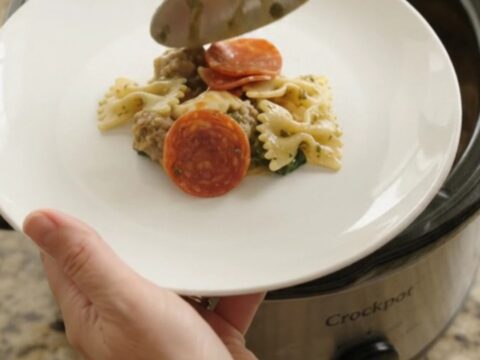 Slow cooker filled with cheesy pepperoni sausage pesto farfalle, with melted cheese and steam rising
