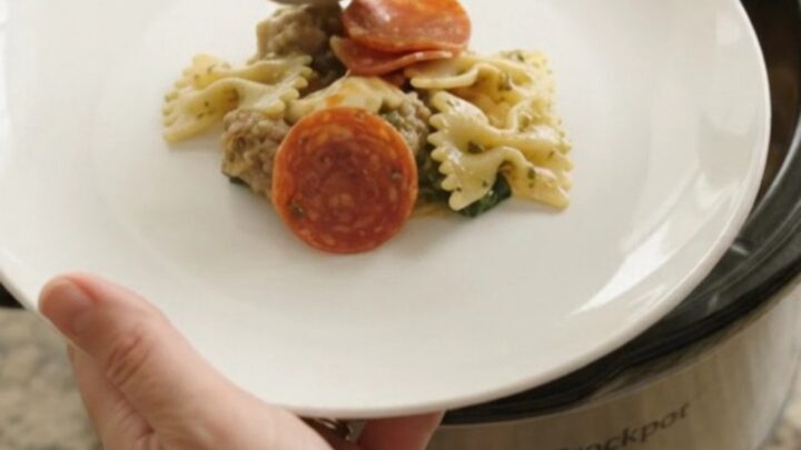 Slow cooker filled with cheesy pepperoni sausage pesto farfalle, with melted cheese and steam rising