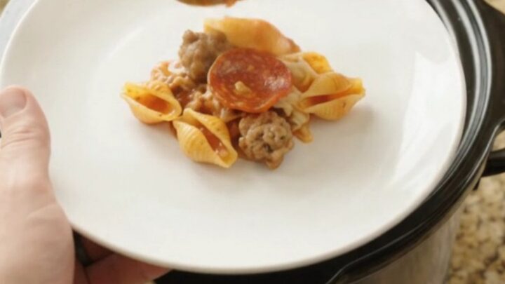 A slow cooker filled with bubbling pasta shells, pepperoni, and sausage in a creamy red pepper sauce