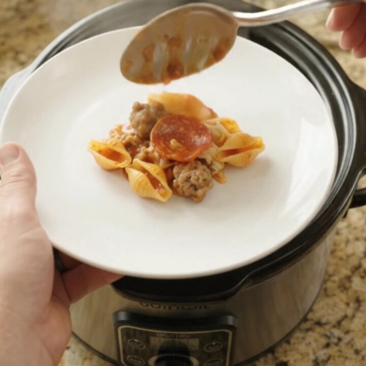 A slow cooker filled with bubbling pasta shells, pepperoni, and sausage in a creamy red pepper sauce
