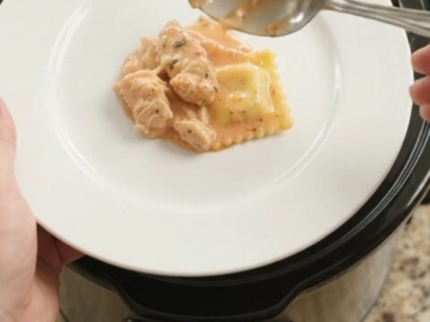 Slow cooker filled with creamy chicken vodka ravioli, topped with melted provolone and ricotta, garnished with basil.