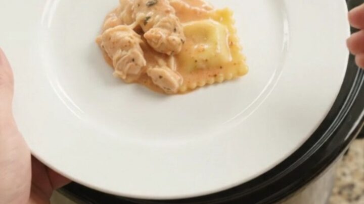 Slow cooker filled with creamy chicken vodka ravioli, topped with melted provolone and ricotta, garnished with basil.
