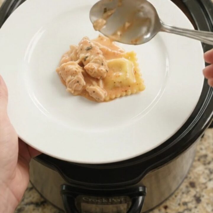 Slow cooker filled with creamy chicken vodka ravioli, topped with melted provolone and ricotta, garnished with basil.