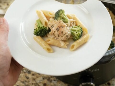 Creamy chicken Alfredo penne with broccoli and melted cheese in a black slow cooker, topped with golden Parmesan crust.
