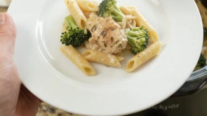 Creamy chicken Alfredo penne with broccoli and melted cheese in a black slow cooker, topped with golden Parmesan crust.