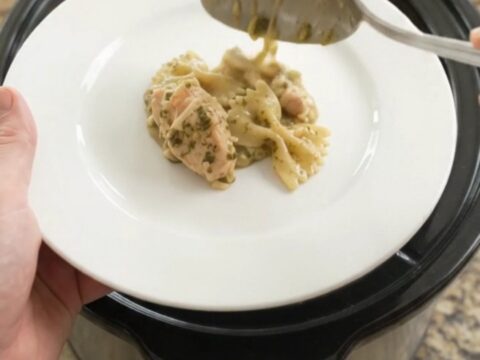 Slow cooker chicken pesto farfalle with melted cheese and spinach, served in a black crockpot.
