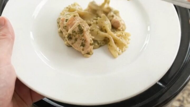 Slow cooker chicken pesto farfalle with melted cheese and spinach, served in a black crockpot.