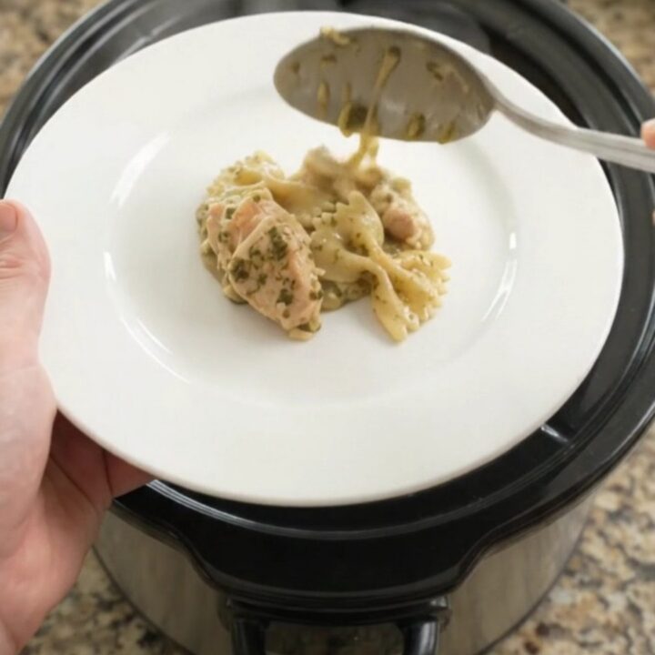 Slow cooker chicken pesto farfalle with melted cheese and spinach, served in a black crockpot.
