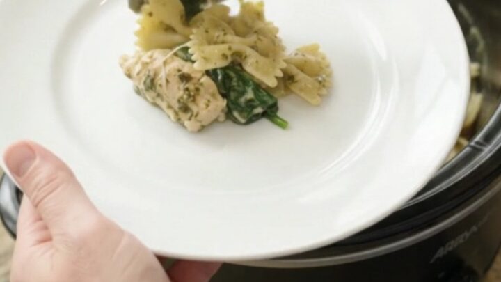 Creamy chicken pesto farfalle in a black slow cooker, with steam rising and melted cheese stretching from a spoon.