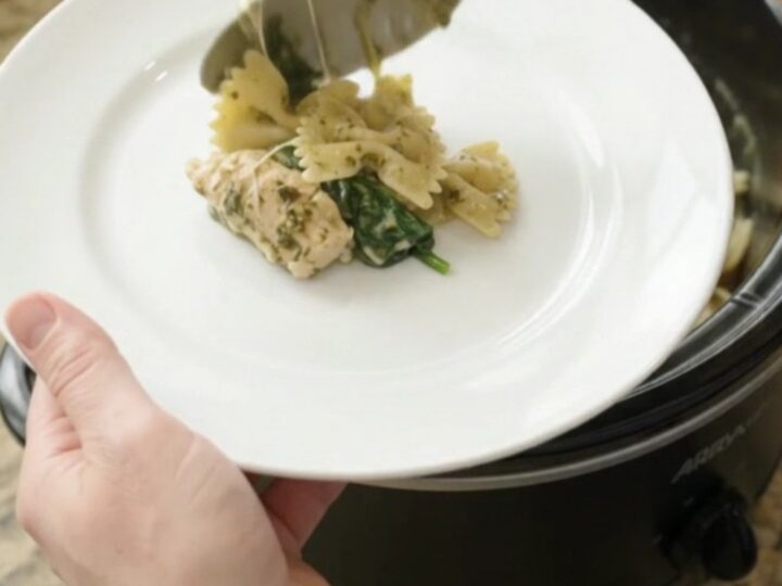 Creamy chicken pesto farfalle in a black slow cooker, with steam rising and melted cheese stretching from a spoon.