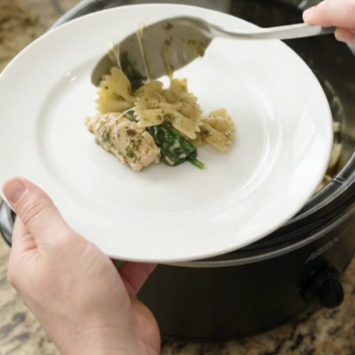 Creamy chicken pesto farfalle in a black slow cooker, with steam rising and melted cheese stretching from a spoon.