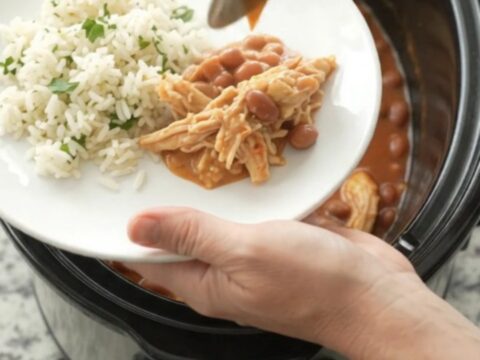 Shredded chicken in creamy red enchilada sauce with melted cheddar, served in a slow cooker with a spoon lifting a bite.